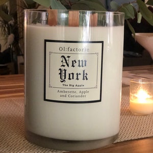 May include: A large, cylindrical candle in a clear glass container. The candle is white with wooden wicks and a label that reads "Ol:factorie New York The Big Apple Amberette, Apple and Coriander."