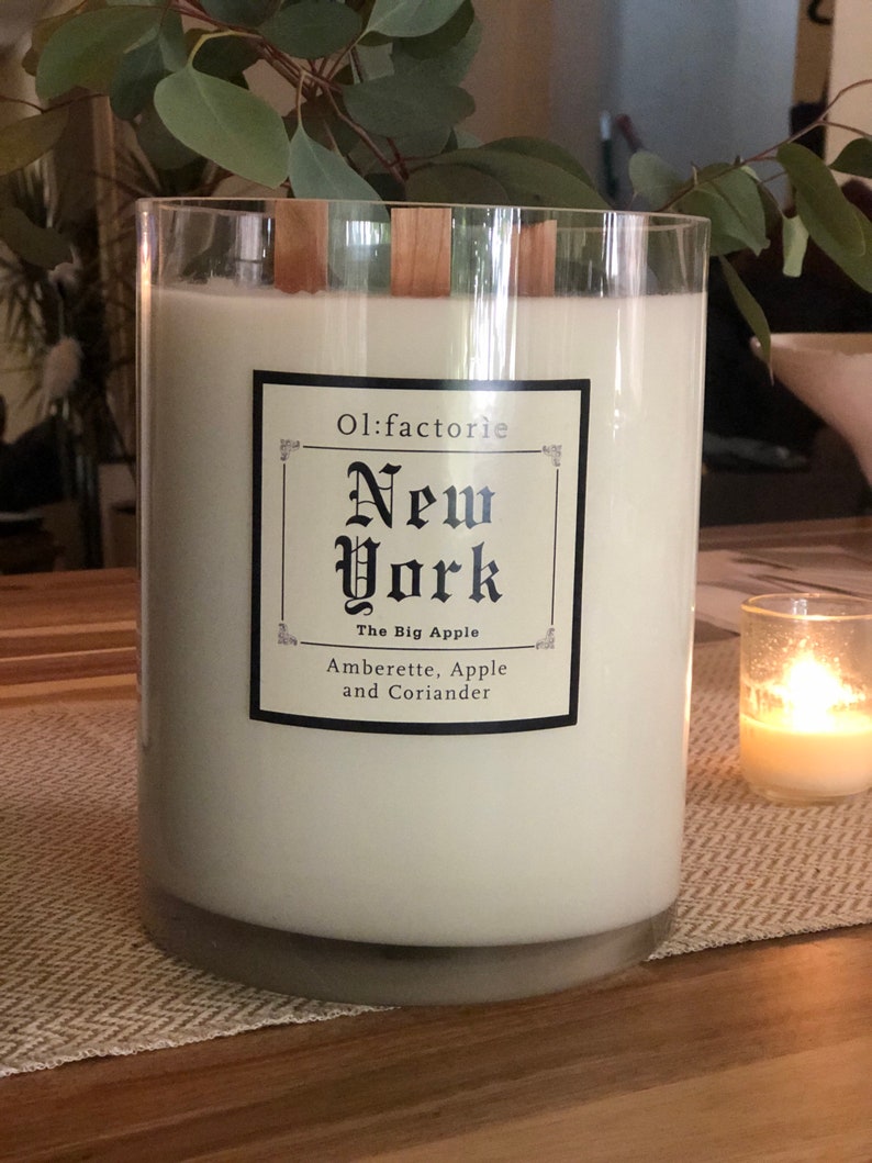 May include: A large, cylindrical candle in a clear glass container. The candle is white with wooden wicks and a label that reads "Ol:factorie New York The Big Apple Amberette, Apple and Coriander."