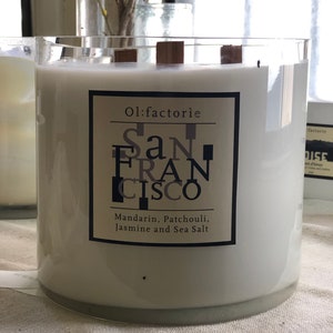 May include: A large, white candle in a clear glass container with wooden wicks. The label reads "Ol:factorie San Francisco" with scent notes of Mandarin, Patchouli, Jasmine, and Sea Salt. The candle is a home decor item.