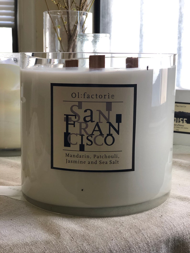 May include: A large, white candle in a clear glass container with wooden wicks. The label reads "Ol:factorie San Francisco" with scent notes of Mandarin, Patchouli, Jasmine, and Sea Salt. The candle is a home decor item.