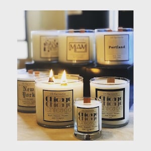 May include: A collection of lit, white candles in glass jars with wooden wicks. Labels include "Chicago", "New York", "Portland", and "MAM". The candles are displayed on a light surface.
