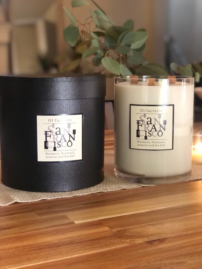 May include: Two candles with the text "San Francisco" are displayed. One is in a black cylindrical container, the other in a clear glass. Both have labels that read "Mandarin, Patchouli, Jasmine and Sea Salt."