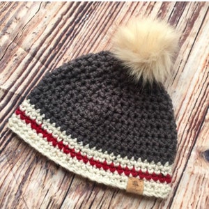 May include: Gray crocheted beanie with a white and red trim and a faux fur pom pom on top. The beanie has a brown leather tag with the word "Handmade" on it.