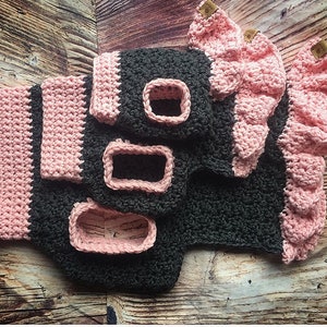 May include: A dark gray crocheted garment featuring light pink trim, ruffled edges, and rectangular openings. The piece includes small brown rectangular tags attached to the pink ruffles, showcasing a textured yarn construction throughout the item.
