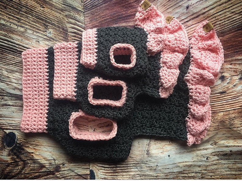 May include: A dark gray crocheted garment featuring light pink trim, ruffled edges, and rectangular openings. The piece includes small brown rectangular tags attached to the pink ruffles, showcasing a textured yarn construction throughout the item.