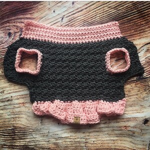 May include: A crochet pet sweater featuring a dark charcoal gray body with a textured stitch pattern. The garment includes a light pink ribbed collar, matching pink trim around the two arm openings, and a ruffled light pink hem at the bottom.