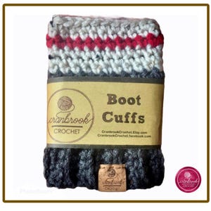 May include: A pair of crocheted boot cuffs featuring a dark gray ribbed base, a light gray middle section, and a red stripe near the top. A tan paper band wraps around the center with the text Cranbrook Crochet Boot Cuffs and social links.