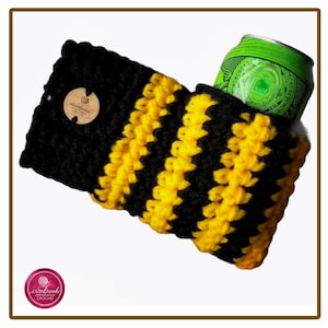 Op de afbeelding: Een zwart-geel gestreepte gehaakte drankhoes met een getextureerd breipatroon. Een rond houten label met de tekst "cranbrook crochet" is aan de zijkant bevestigd. Een groen aluminium drankblikje is in de hoes gestoken.