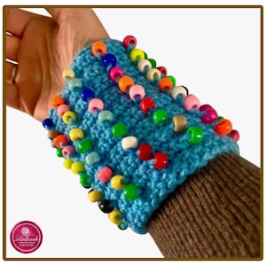 Könnte beinhalten: Eine hellblaue, gehäkelte Armstulpe mit Reihen aus bunten Kunststoffperlen in den Farben Rot, Orange, Gelb, Grün, Rosa und Weiß. Das strukturierte Strickaccessoire wird am Unterarm getragen und zeigt ein lebendiges, mehrfarbiges Muster.