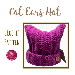 Puede incluir: Un gorro de punto de color morado con orejas de gato. El gorro está hecho con un punto texturizado y tiene una pequeña etiqueta marrón en el lateral.