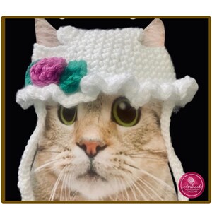 May include: A white crocheted pet hat featuring a ruffled brim and two decorative yarn flowers in magenta and teal. The hat has long side ties and is displayed on a cat. A small circular logo in the bottom right corner reads cranbrook CROCHET.