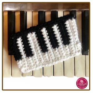 Può includere: Una fascia rettangolare all'uncinetto con un motivo a tasti di pianoforte bianco e nero. Il design è composto da punti di filato alternati bianchi e neri disposti per imitare l'aspetto dei tasti di una tastiera musicale, con un bordo superiore nero.