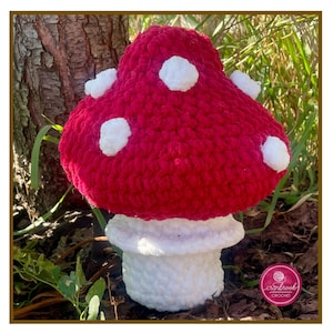 May include: A handmade, red crochet mushroom with white spots and a white stem. The mushroom is displayed outdoors near a tree trunk and greenery. The mushroom is a whimsical decorative item.