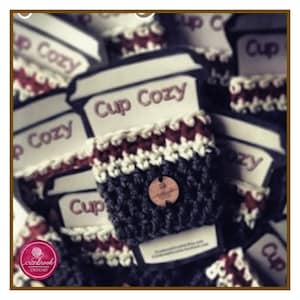 Könnte beinhalten: Nahaufnahme eines gehäkelten Becherwärmer in Schwarz, Weiß und Burgunderrot. Der Wärmer hat einen Knopf aus Holz und ein Etikett mit der Aufschrift "Cup Cozy".