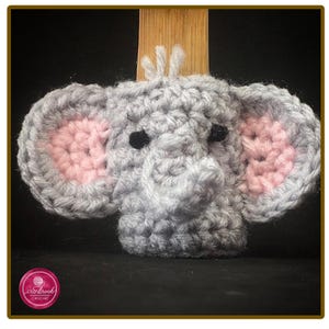 May include: A grey crocheted elephant head with large ears featuring pink centres, black yarn eyes, and a small trunk. The elephant has a tuft of grey yarn on top of its head and is crafted with a textured stitch pattern throughout the design.