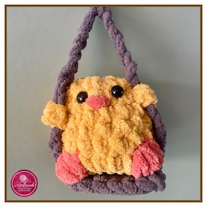 May include: A crocheted yellow chick with pink feet and black eyes is sitting in a purple crocheted swing. The swing has a purple crocheted strap.
