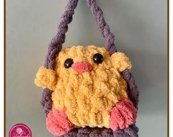 May include: A crocheted yellow chick with pink feet and black eyes is sitting in a purple crocheted swing. The swing has a purple crocheted strap.