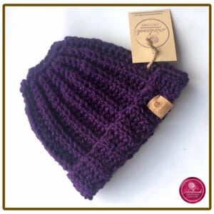 Può includere: Un berretto all'uncinetto color viola scuro con una trama a coste e un bordo ripiegato. Un'etichetta rettangolare color cuoio con il testo Cranbrook Crochet è fissata sul lato, e un'etichetta di carta più grande è legata in cima con uno spago.