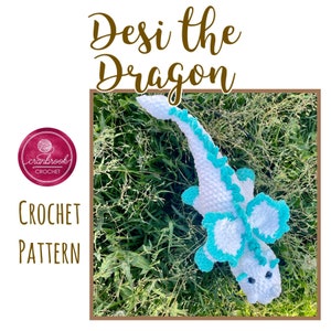 May include: Crochet pattern for a white and turquoise dragon amigurumi toy. The dragon is lying on a bed of green grass. The pattern is called "Desi the Dragon" and is by Cranbrook Crochet.
