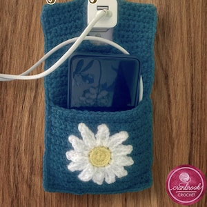 May include: Teal crochet charging station with a white daisy applique. The station holds a phone and charger, with a white cord extending from the top. The design includes a pocket for the phone and a plug-in for the charger.