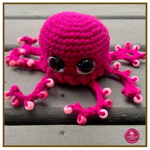 May include: A handmade, magenta crochet octopus with large, black, glittery eyes. The octopus has eight tentacles, each adorned with pink beads. The crochet work is detailed, and the toy is set against a neutral background.