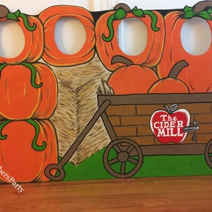 May include: A painted cardboard photo booth prop featuring a fall theme with pumpkins, hay bales, and a wagon with the text "The Cider Mill".