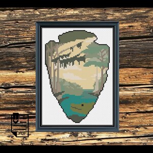 May include: A cross-stitch pattern of a National Park Service arrowhead design. The design features a green and brown forest scene with a blue lake and a small island in the center.