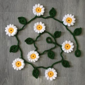 May include: A crocheted daisy garland with white petals and yellow centers, connected by green stems and leaves. The handmade floral decoration is arranged on a gray wooden surface.
