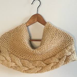 May include: A tan-colored knitted cowl scarf with a braided design along the bottom edge. The scarf is displayed on a wooden hanger against a white wall. The knitted texture is visible.