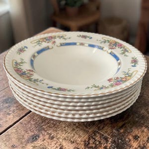 May include: A stack of white ceramic soup bowls with a scalloped edge and a floral and blue border design. The bowls have a gold trim and are stacked on a wooden surface.