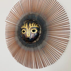 May include: A metal sunburst wall art piece. The central face is a dark, textured metal with gold accents. Copper-coloured rods radiate outwards, creating a sun-like design. The artwork is round and appears to be mounted on a white wall.