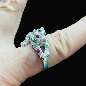May include: A silver ring with a leopard design. The leopard is decorated with clear crystals and red, green, and blue gemstones. The ring is set against a black background.