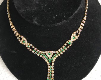 May include: A gold-toned necklace with clear and green rhinestones. The necklace features a Y-shaped pendant with dangling green rhinestones. The necklace is displayed on a black velvet bust.