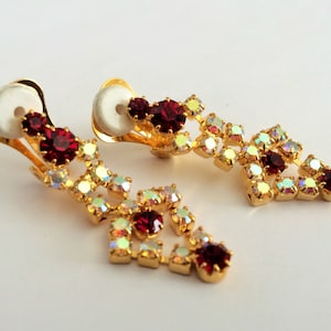 May include: A pair of gold-tone clip-on earrings featuring red and iridescent clear rhinestones. The earrings are shaped like teardrops.