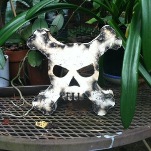 May include: A white metal skull and crossbones sculpture with a distressed finish. The sculpture is sitting on a metal table with a grid pattern. The sculpture is surrounded by green plants.