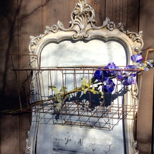 May include: A vintage-style wall-mounted basket with a distressed, ornate frame. The basket is made of rusty metal and holds purple and yellow faux flowers. The frame is painted in a distressed white and gray color scheme.