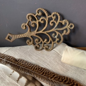 May include: A gold-toned metal trivet in the shape of a stylized tree with intricate details. The trivet has a square handle on one end.