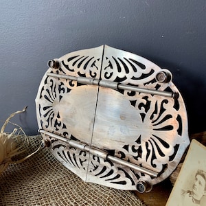 May include: A silver-plated, oval-shaped, decorative, folding,  plate stand with intricate cut-out designs. The stand has a hinged design and is made of metal.