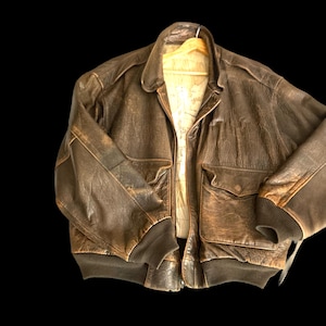 May include: A dark brown leather bomber jacket featuring a distressed texture, a wide collar, long sleeves with ribbed cuffs, a ribbed hem, and two large front pockets with flaps. The interior lining displays a light-colored map print pattern.