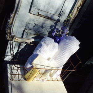 May include: A rustic wire basket holds white towels, soap, and a small bouquet of purple flowers. The basket sits on a white surface, with a distressed, light blue and white painted door frame in the background.