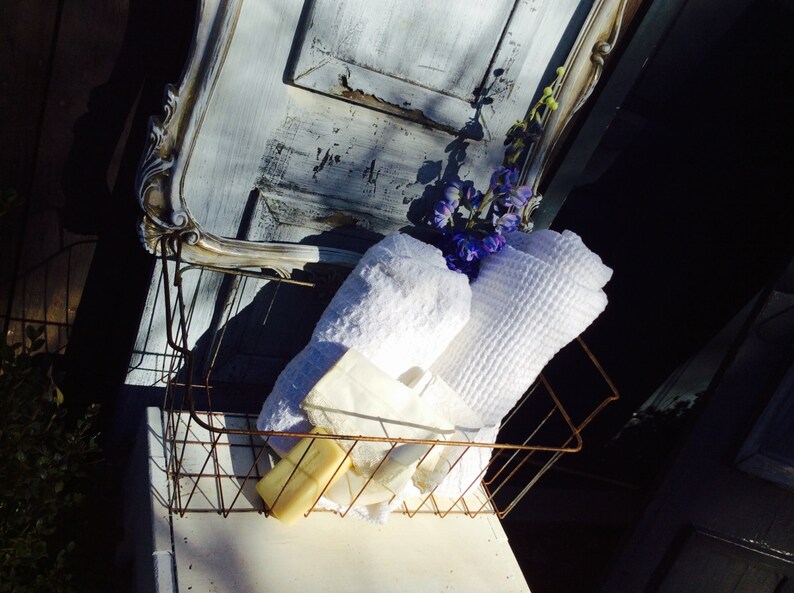 May include: A rustic wire basket holds white towels, soap, and a small bouquet of purple flowers. The basket sits on a white surface, with a distressed, light blue and white painted door frame in the background.