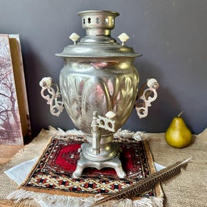 May include: A metallic silver samovar featuring a bulbous body with vertical fluting, ornate side handles, and a central spigot. The vessel includes white knob accents on the lid, handles, and tap, set atop a four-legged base with a chimney top.