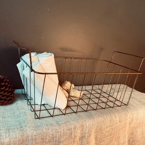 May include: A rectangular, rusty brown wire basket with handles, containing rolled white towels and seashells. The basket sits on a textured, light-colored surface. A pinecone is visible to the left of the basket.