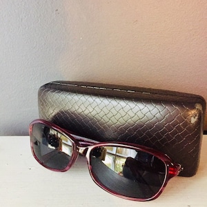May include: Burgundy-colored sunglasses with dark lenses and a black case. The sunglasses have a rounded, oval shape.