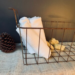 May include: A rustic, wire basket with a handle, holding folded white towels and several natural sponges. A large pine cone sits to the left. The basket has a weathered, brown finish, suggesting a vintage or farmhouse aesthetic.