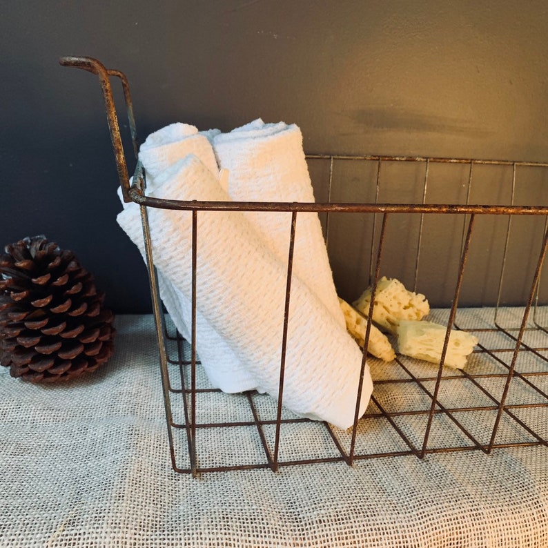 May include: A rustic, wire basket with a handle, holding folded white towels and several natural sponges. A large pine cone sits to the left. The basket has a weathered, brown finish, suggesting a vintage or farmhouse aesthetic.