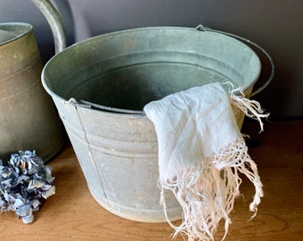May include: A vintage galvanized metal bucket with a white fabric with fringe hanging over the edge. The bucket is sitting on a wooden surface.