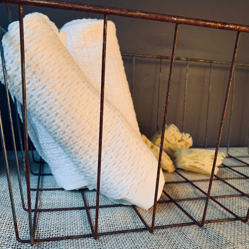 May include: A rustic, wire basket with rolled white towels and several beige sponges. The basket has a distressed, brown finish and sits on a textured, neutral-colored surface. The towels are neatly folded, and the sponges are irregularly shaped.