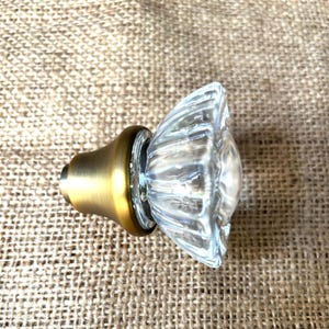 May include: A clear glass door knob with a ribbed design and a brass-colored base. The knob is a classic design, suitable for interior doors. The glass is transparent, allowing light to pass through.