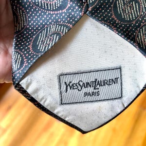 May include: A dark necktie featuring a repeating pattern of circular motifs in pink and white dots. The back of the tie has a cream-colored lining with a rectangular black and white label that reads Yves Saint Laurent Paris in a serif font.
